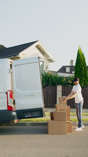 A young man wearing a white T-shirt and jeans is shown standing next to a large white removal van with its side door open, engaged in loading or unloading packed cardboard boxes. The boxes, variations in sizes and shapes, are stacked on the pavement beside him, and he is holding one box in his hands, preparing to place it inside or remove it from the van. The scene is set outdoors on a suburban street with residential houses and a wooden fence in the background,, and a tall green shrub visible near the fence. The van is positioned near the curb on a clear, sunny day, with natural daylight illuminating the scene. This image illustrates the process of home relocation, packing and moving, as the individual manages furniture transport and loading operations, with the interior space of the van ready to accommodate more items. The setting is typical for professional removals, representing a structured loading process coordinated by Removal Van Kennington as part of their removals services.