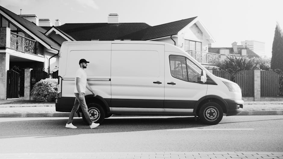 A man with a beard, wearing a baseball cap, white t-shirt, and light-colored jeans, is walking on a paved street beside a large white panel van with black detailing in the vicinity of a residential area. The van is parked close to the pavement outside a house with a modern design, featuring a balcony with a railing, large windows, and a sloped roof. The van's sliding door appears to be closed, and the vehicle is positioned parallel to the house, suggesting a home relocation or furniture transport process. In the background, there are other houses with similar architectural styles, gardens, and fences, with some greenery visible. The street is clean, and the scene is captured during daylight with clear lighting. This image exemplifies the typical loading or moving process managed by Removal Van Kennington as part of professional removals and packing and moving services, with equipment like trolleys or blankets possibly used but not visible in this shot.