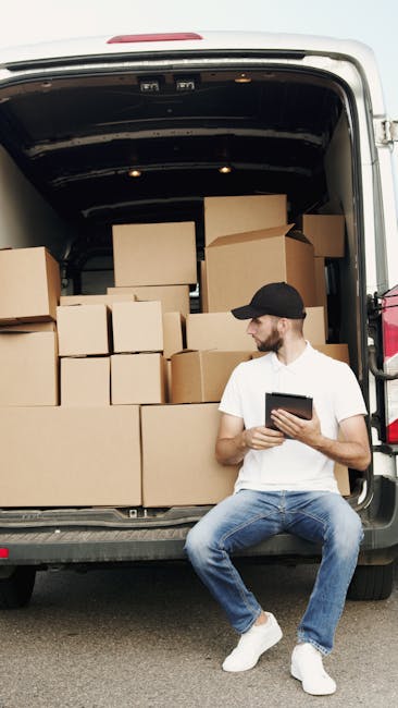 A young man wearing a white T-shirt and jeans is shown standing next to a large white removal van with its side door open, engaged in loading or unloading packed cardboard boxes. The boxes, variations in sizes and shapes, are stacked on the pavement beside him, and he is holding one box in his hands, preparing to place it inside or remove it from the van. The scene is set outdoors on a suburban street with residential houses and a wooden fence in the background,, and a tall green shrub visible near the fence. The van is positioned near the curb on a clear, sunny day, with natural daylight illuminating the scene. This image illustrates the process of home relocation, packing and moving, as the individual manages furniture transport and loading operations, with the interior space of the van ready to accommodate more items. The setting is typical for professional removals, representing a structured loading process coordinated by Removal Van Kennington as part of their removals services.