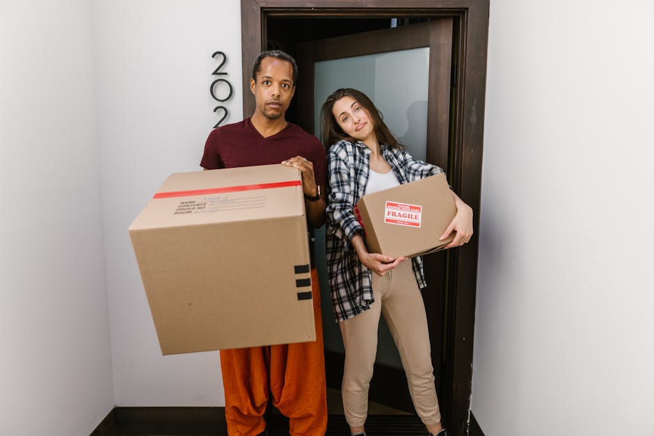 A man and a woman stand side by side inside a hallway near a doorway, each holding a cardboard box used for home relocation. The man, on the left, has a dark complexion, short dark hair, and is dressed in an orange pair of trousers and a dark maroon T-shirt. The woman, on the right, has a fair complexion, long brown hair, and is wearing a checked shirt over a white T-shirt with beige trousers. The woman leans her head slightly towards the man, who looks directly at the camera with a serious expression. The woman holds a medium-sized cardboard box labeled 'Fragile' with red tape, while the man supports a larger cardboard box with black tape and shipping labels. The boxes are made of brown cardboard, and the woman appears to be in the midst of a packing and moving process, as part of a house removal effort. The background reveals a dark wooden door frame and a white wall, with a glimpse of a temporary environment for furniture transport and loading. This scene reflects the logistics involved in professional removals, where packing, carrying, and transporting household items are key elements supported by companies like Removal Van Kennington, dedicated to efficient house removals.