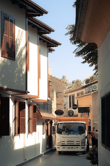 A narrow street between two two-storey buildings with overhanging roofs and wooden shutters on the windows. In the middle of the scene, a white moving van with the logo of Removal Van Kennington is parked, partially blocking the passage. The van is positioned close to the building on the left, with its front facing towards the camera. The street appears to be used for home relocation activities, with the van likely being part of a furniture transport or packing and moving process. The surroundings include a mix of residential and commercial signs on the building in the background, and a person is visible to the right side near the van, possibly involved in the loading process. This setting suggests an active moving site during daylight hours, with items to be loaded or unloaded, supporting a house removal or furniture transport service context as provided by [COMPANY_NAME].