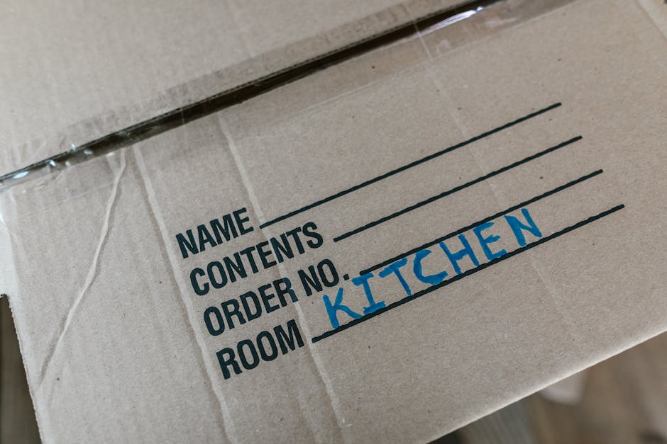 Close-up image of a brown cardboard moving box with black printed labels for 'Name', 'Contents', 'Order No.', and 'Room' followed by handwritten text in blue ink indicating 'KITCHEN'. The box appears