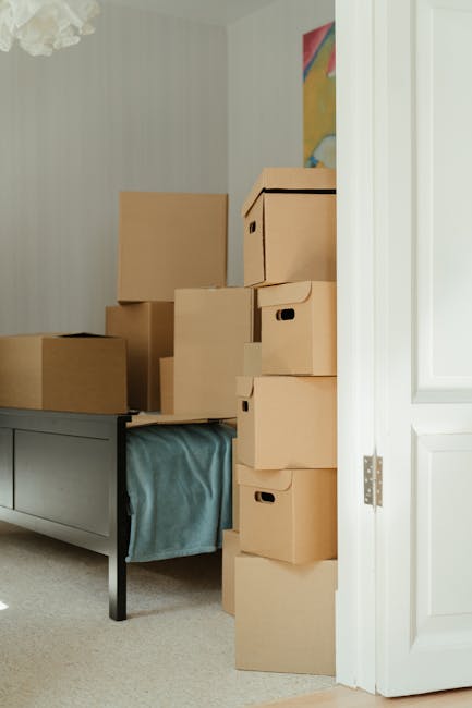 Inside a home room, several brown cardboard boxes of varying sizes are stacked on top of a grey furniture piece with a blue fabric cover. The boxes are arranged near an open white door, with some boxe