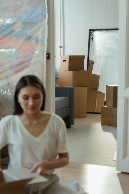 Inside a home room, several brown cardboard boxes of varying sizes are stacked on top of a grey furniture piece with a blue fabric cover. The boxes are arranged near an open white door, with some boxe