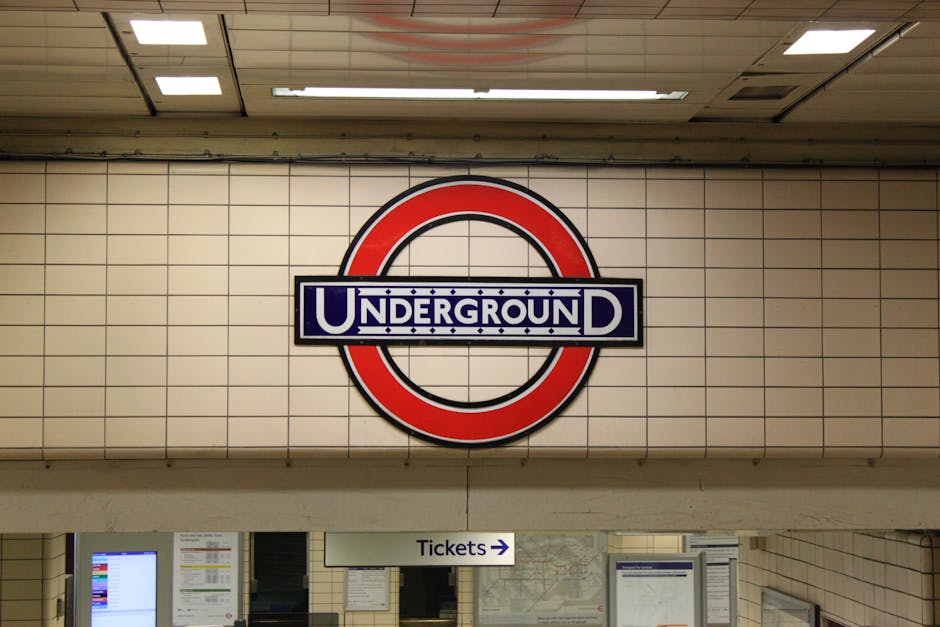 Close-up of the London Underground roundel sign with a red circle and blue bar displaying the word 'UNDERGROUND' in white capital letters, mounted on a beige tiled wall near the station ticket area. Below the sign, a small white arrow points to the right, indicating the ticketing direction, with a sign reading 'Tickets' visible beneath. The station environment is well-lit with ceiling lights, and the surrounding area includes ticket machines and informational displays, reflecting typical station infrastructure associated with home relocation and transport logistics services offered by Removal Van Kennington in the context of house removals and moving routes.