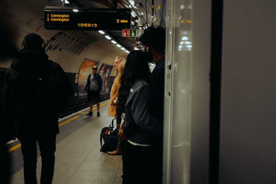 Photograph taken inside Kennington tube station showing multiple commuters waiting on the platform, with some standing near the edge and others further back. A digital display overhead indicates upcoming train routes and times, including a train to Kennington scheduled in five minutes. The station's tunnel features curved walls with overhead lighting illuminating the area. Several individuals are wearing casual clothing and carrying backpacks or small bags, reflecting typical passengers on a daily commute. The platform is clean, with a textured surface, and the background includes the subway tracks and a partially visible train door. This setting captures a moment of home relocation or transport planning, relevant to house removals and moving services provided by Removal Van Kennington, demonstrating the typical environment where furniture transport and moving logistics are coordinated.