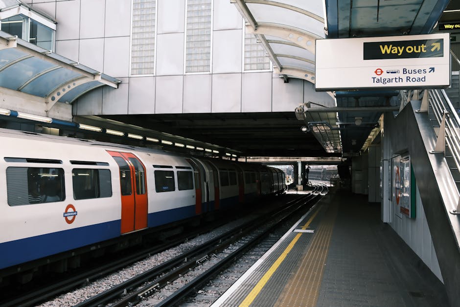 Close-up of the London Underground roundel sign with a red circle and blue bar displaying the word 'UNDERGROUND' in white capital letters, mounted on a beige tiled wall near the station ticket area. Below the sign, a small white arrow points to the right, indicating the ticketing direction, with a sign reading 'Tickets' visible beneath. The station environment is well-lit with ceiling lights, and the surrounding area includes ticket machines and informational displays, reflecting typical station infrastructure associated with home relocation and transport logistics services offered by Removal Van Kennington in the context of house removals and moving routes.