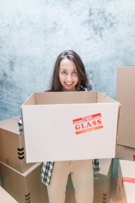 A woman with long brown hair smiling and standing indoors in front of a pale blue wall, holding an open cardboard box labeled 'GLASS' in red print. She is dressed casually in a plaid shirt and light-colored trousers. Around her are several other packed cardboard boxes of various sizes, some sealed with packing tape, positioned on the floor and stacked nearby. The scene depicts a packing or home relocation process, with the boxes ready to be loaded onto a removal van. The environment suggests an interior space prepared for a furniture transport or moving service, with bright, even lighting highlighting the packing materials and assisting in the loading process, as done by a professional moving company like Removal Van Kennington.
