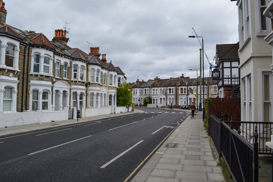 A wide view of a residential street in Kennington on an overcast day, showing a row of Victorian-style terraced houses with white facades, bay windows, and brick chimneys along the left side of the road. The pavement on the right side features a black metal fence and a few potted plants beside a cream-colored building. The street is relatively quiet, with only a few pedestrians visible in the distance, and the road has clear lane markings. In the background, construction cranes and scaffolding indicate ongoing building work. The image captures the typical urban environment suitable for house removals and furniture transport services, as provided by Removal Van Kennington, supporting efficient packing and loading processes in a residential area.