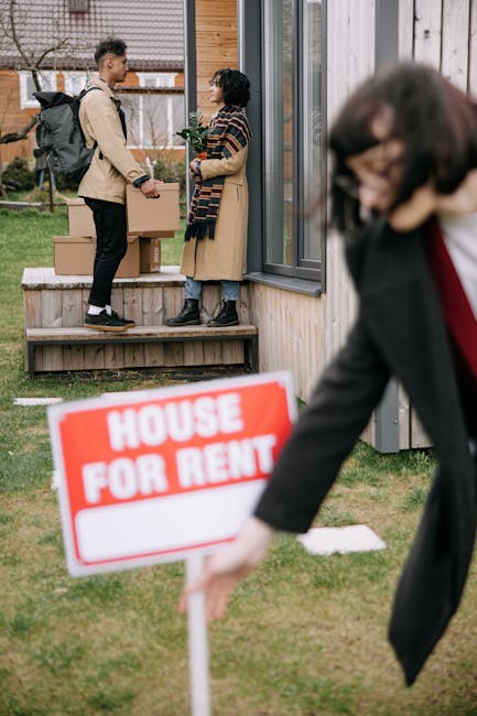 A woman with long brown hair smiling and standing indoors in front of a pale blue wall, holding an open cardboard box labeled 'GLASS' in red print. She is dressed casually in a plaid shirt and light-colored trousers. Around her are several other packed cardboard boxes of various sizes, some sealed with packing tape, positioned on the floor and stacked nearby. The scene depicts a packing or home relocation process, with the boxes ready to be loaded onto a removal van. The environment suggests an interior space prepared for a furniture transport or moving service, with bright, even lighting highlighting the packing materials and assisting in the loading process, as done by a professional moving company like Removal Van Kennington.
