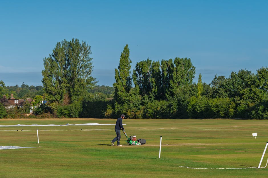 A person using a lawn mower on a large, well-maintained grassy area with neatly trimmed lawn and clearly marked boundary markers made of white posts and thin cords. In the background, there are tall, leafy green trees and residential houses situated beyond the open space, under a bright blue sky with minimal cloud cover. The scene is outdoors, during daylight hours, and the individual appears engaged in turf maintenance, possibly related to preparing a sports field or open recreational area. The setting is peaceful and organized, with the spacious surroundings showcasing a community park or sports ground, which could be linked to local amenities near residential areas such as those around Oval cricket ground. The presence of the mower and the maintained grass suggests ongoing landscaping or groundskeeping, relevant to property management or outdoor space care, which may be coordinated by services like Removal Van Kennington for house or property relocations involving outdoor space preparation.