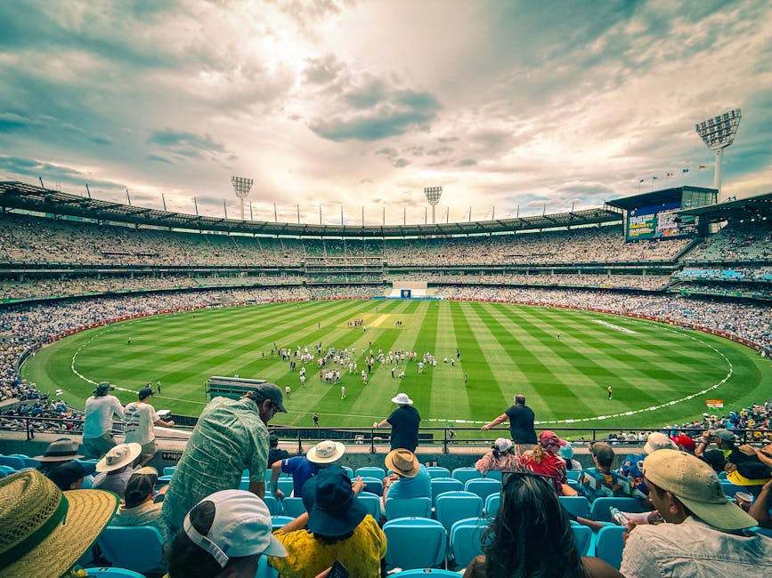 A panoramic view of a large cricket stadium with a well-maintained grass pitch and clearly marked boundary lines. The stadium features multiple levels of seating filled with spectators, many wearing hats and casual clothing, seated on blue plastic chairs. Several individuals are standing and observing the field, while others are seated. The sky above the stadium is partly cloudy with sunlight filtering through, creating a bright and open atmosphere. In the foreground, some spectators are leaning over the railing, watching the field and the ongoing activities on the pitch. The stadium's floodlights are mounted on tall structures, though they are not illuminated. This scene illustrates the scale of a sports venue, with attention to the seating arrangements and environment, similar to the context of planning a home relocation or physical transport process with a professional removal service such as Removal Van Kennington, supporting the themes of logistics and organized movement.