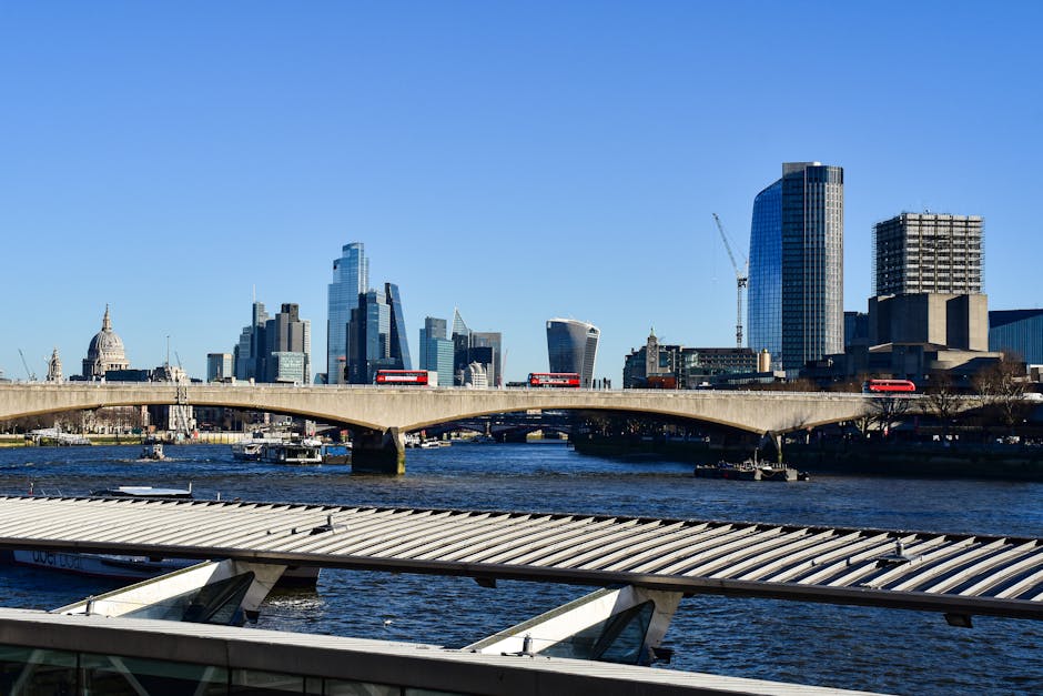 A wide view of the London city skyline across the River Thames on a clear day, featuring modern high-rise buildings including glass office towers and cranes indicating ongoing construction. In the foreground, a large metallic and glass-roofed structure extends over part of the river, while a concrete bridge with red double-decker buses transports vehicles across the water. The scene captures the urban environment typical of a busy city with tall skyscrapers, some with reflective glass facades, and the iconic St. Paul's Cathedral dome visible in the distance. The water is calm, with a few boats floating near the banks, and the sky is bright blue with no clouds, highlighting the vibrant, active presence of London’s financial and commercial district. This setting aligns with house removals and relocation services in the area, illustrating the city environment where professional furniture transport and home relocation are commonly coordinated through companies like Removal Van Kennington.
